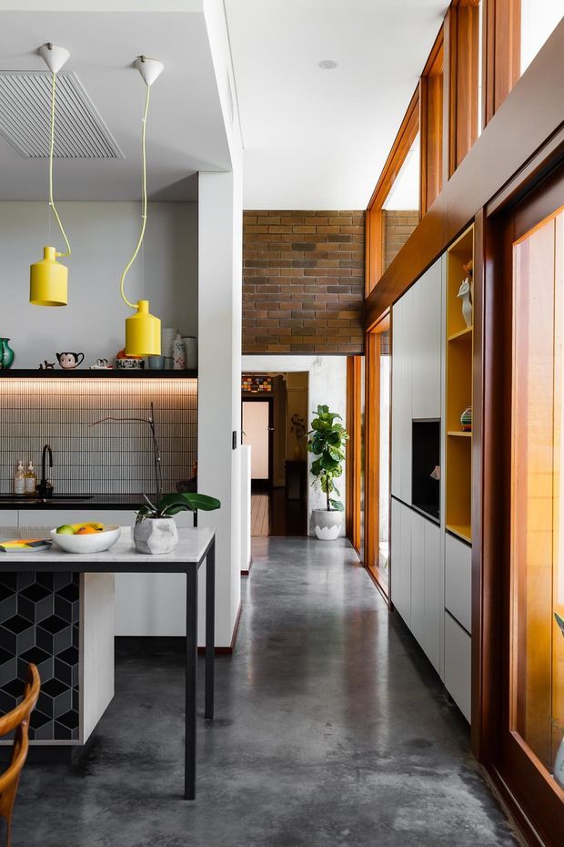 A bright, contemporary open-plan kitchen and hallway with polished concrete flooring, white walls and cabinetry, a black geometric-tile island face, and a dark ribbed-tile backsplash. Yellow pendant lights add a bold accent above the kitchen bench. The adjoining circulation space features full-height timber-framed glazing, exposed brickwork, built-in display shelving, and integrated media cabinetry.