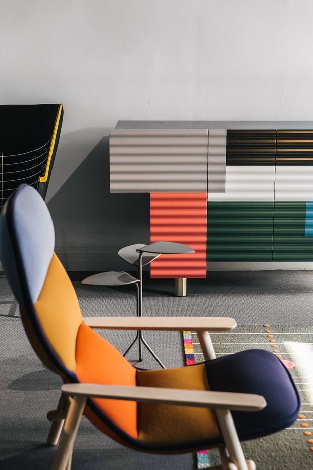 A styled interior vignette featuring a colourful mid-century-inspired lounge chair in the foreground, with pale timber arms and legs, dark blue and orange upholstery, and a black frame. Behind it is a low sculptural sideboard or storage unit with horizontally ribbed panels in grey, white, red, and deep green, accompanied by small sculptural side tables. The space has a grey carpet, white textured wall, and strong natural sunlight creating dramatic shadows.