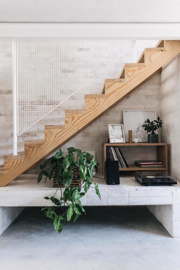 Interior view of a compact industrial-style living area beneath a staircase. A pale plywood stair with open stepped detailing runs along a white-painted brick wall, paired with a white metal balustrade. Below, a built-in white brick bench or ledge supports a dark timber media console with records, framed art, a turntable and a speaker. Green indoor plants add a natural accent, and the floor is smooth grey concrete.