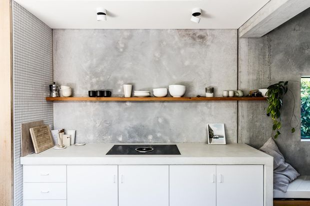 A minimalist kitchen or kitchenette wall featuring white flat-panel base cabinetry, a pale stone or concrete benchtop with an integrated black cooktop, a long timber display shelf, and a mottled grey concrete-look wall finish. The left wall is clad in small-format light tiles, while open shelving displays ceramics and kitchenware. A leafy plant and soft seating are visible at the right edge.