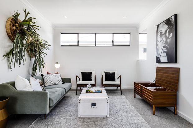Bright, refined living room with a grey sofa, two mid-century-style armchairs, a white storage coffee table, a textured grey rug, and a wooden record console. White walls and ceiling, high horizontal windows with dark frames, a sculptural wall-mounted plant, and monochrome artwork complete the space.