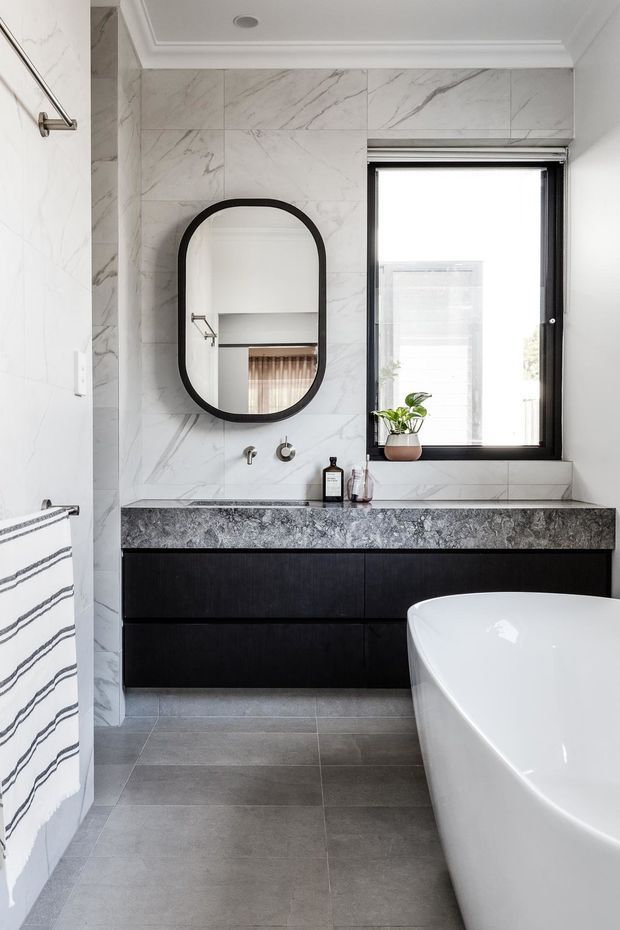 Bright contemporary bathroom featuring large marble-look wall tiles, grey floor tiles, a black timber-look floating vanity with a dark stone benchtop, black-framed window, arched black-framed mirror, and a white freestanding bathtub. The space has crisp white walls and ceiling, strong natural light, and restrained monochrome styling.