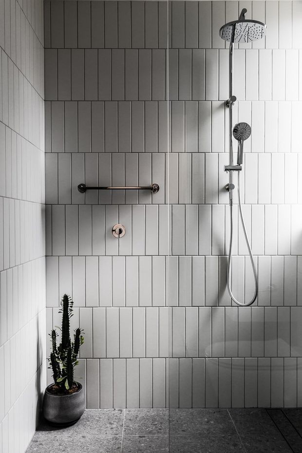 A minimalist walk-in shower with vertically oriented light grey rectangular wall tiles arranged in horizontal bands, dark grey stone-look floor tiles, and sleek chrome rain shower and handheld fittings. A black metal grab rail and small potted cactus add functional and decorative details.
