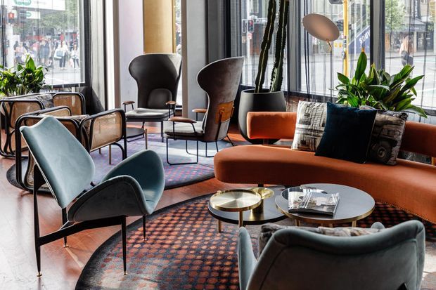 A stylish urban lounge with floor-to-ceiling windows, a curved rust-orange sofa, sculptural upholstered lounge chairs, patterned rugs, round coffee tables, indoor plants, and a warm contemporary palette. City streets and buildings are visible outside the glazing.