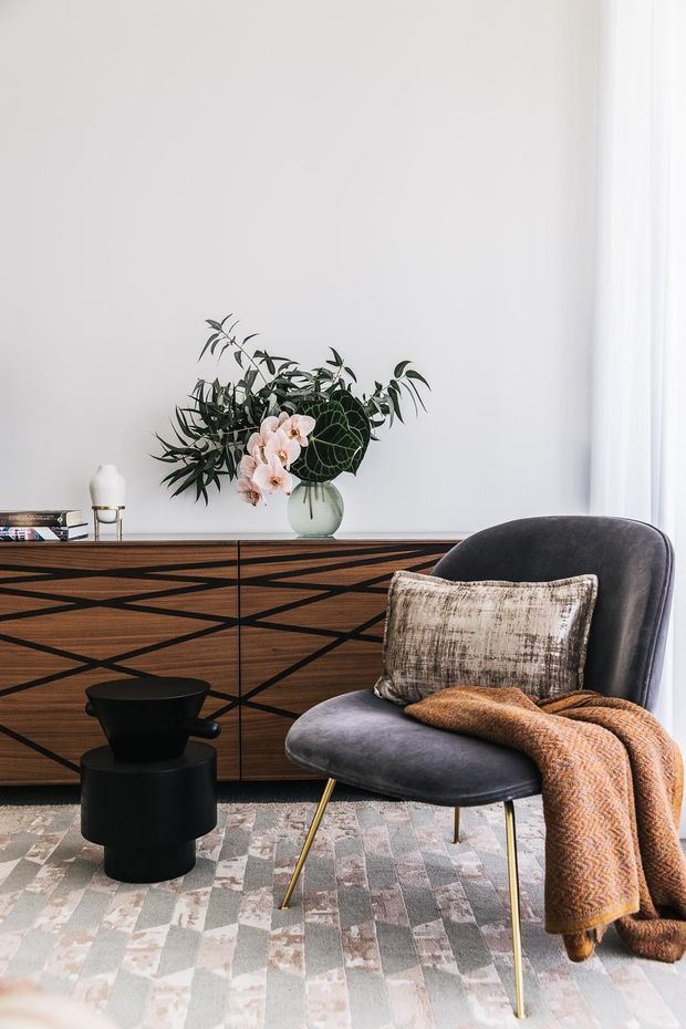 A refined living room vignette with a mid-century-inspired dark timber sideboard featuring geometric black inlay, a charcoal upholstered accent chair with slender brass-toned legs, a textured neutral cushion, and a rust-brown throw. A black sculptural side table, patterned area rug, floral arrangement, and sheer curtains complete the softly lit interior.