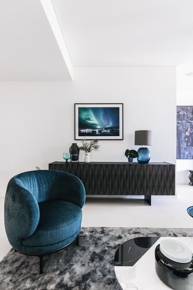 Bright contemporary living room with white walls and tiled flooring, featuring a deep blue velvet lounge chair, a dark geometric-patterned sideboard, blue and black decorative vessels, framed artwork, and a grey abstract rug. A glimpse of an adjacent space is visible at the right edge.