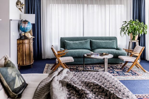 A stylish living room with a teal upholstered sofa, mid-century folding lounge chairs, a round coffee table, layered patterned rugs, and deep blue carpet. Full-height sheer curtains with dark navy drapes frame the room, while a timber cabinet, globe, plants, and decorative objects add warmth and character.