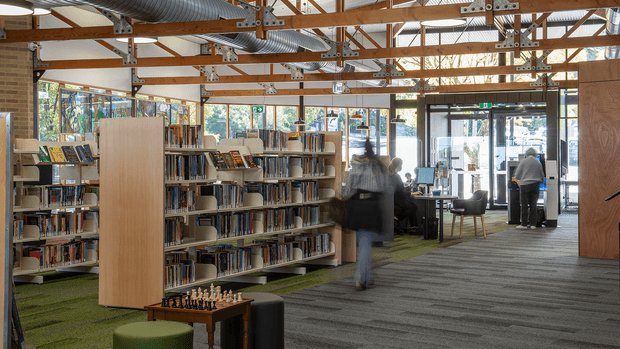 A bright, contemporary library interior with rows of open bookshelves, reading and study tables, upholstered stools and chairs, and a glazed entrance at the far end. The space has exposed timber roof beams, visible metal structural connections, exposed ventilation ductwork, and extensive windows. Green carpet tiles define the shelving area, while grey carpet covers the main circulation zone. A chess table sits in the foreground.