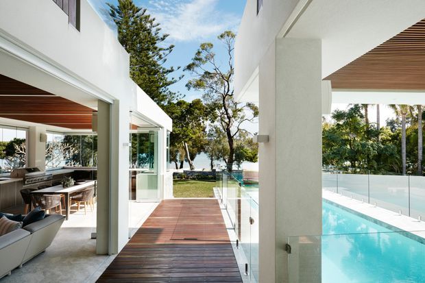 Bright contemporary residential courtyard or terrace view with a timber deck walkway, glass balustrades, white rendered walls, and a turquoise swimming pool. An open-plan dining and living area is visible to the left, while the view extends toward landscaped grounds and trees beyond.