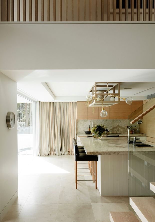 A bright, refined open-plan kitchen viewed from an adjacent circulation area. The space features pale stone flooring, a large light-stone island with seating, vertically reeded pale cabinetry, warm timber wall cabinetry, a stone backsplash, expansive glazing with full-height cream curtains, and a sculptural timber-and-metal pendant light. A stair and upper-level balustrade are visible, suggesting a compact multi-level contemporary home.