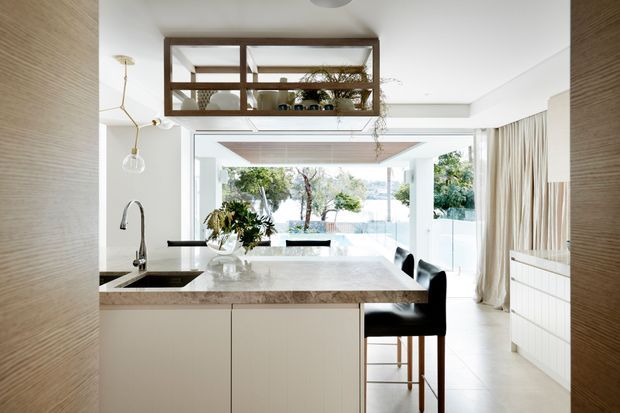 Bright contemporary open-plan kitchen with a large pale stone island and integrated sink, white cabinetry, dark bar stools, and a timber-framed glazed display partition above the kitchen. Floor-to-ceiling glazing and sheer curtains open onto a landscaped outdoor area, creating an airy indoor-outdoor connection. Pendant lighting, concealed storage, and light neutral finishes complete the refined interior.