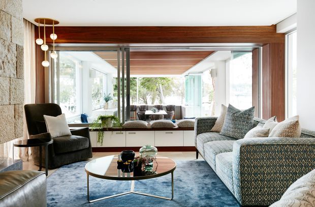 A bright, refined living room with floor-to-ceiling glazing, a large patterned sectional sofa, lounge chair, round coffee table, blue area rug, and a built-in window seat. Rich timber ceiling and wall framing contrast with white walls, while a large mirror visually extends the room and reflects the seating area.