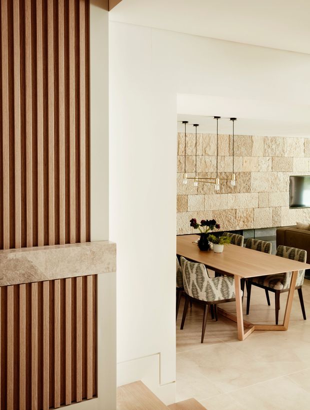 A refined open-plan dining area viewed through a wide interior opening. The space features a warm timber dining table with woven upholstered chairs, a textured light stone feature wall, pendant lighting, pale stone flooring, and a timber-slat feature wall with a built-in stone ledge in the foreground. The palette is warm, neutral, and contemporary.