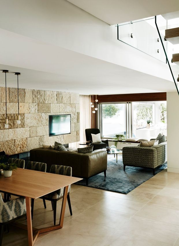 Bright open-plan interior combining a dining area and living room, with a large timber dining table in the foreground, leather and woven lounge seating, a dark blue rug, a textured natural stone feature wall, expansive glazing, and a visible upper-level stair or void with glass balustrading. The space has a clean contemporary architectural shell and warm natural materials.