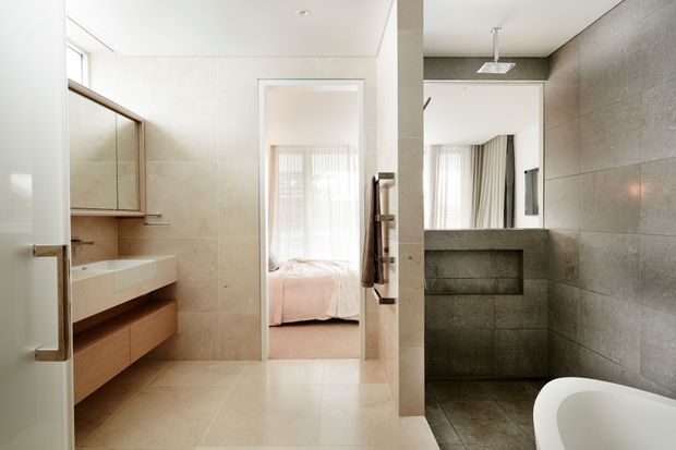 Contemporary bathroom with large beige stone-look wall and floor tiles, a floating timber vanity with integrated white basin, frameless mirror, shower area with darker grey tile finishes, and a freestanding white bathtub in the foreground. An open doorway provides a clear view into an adjacent bedroom.