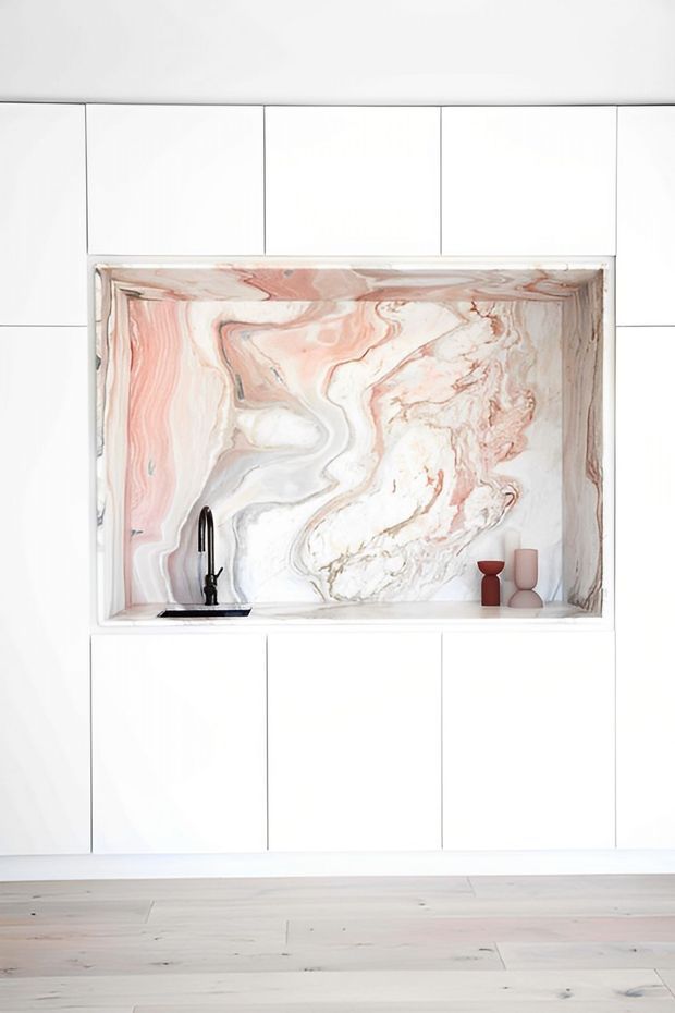 Bright minimalist interior detail featuring a white tiled wall with large rectangular grid tiles, a recessed marble-look feature niche, and a compact black tap and basin. The niche has dramatic flowing pink, white, and grey veining, with small decorative vessels on the ledge. Pale timber flooring is visible below.