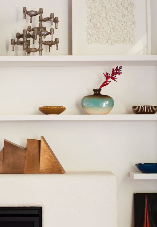 Close-up interior detail of white built-in display shelving above and beside a textured white fireplace. The shelves hold sculptural metal and ceramic decorative objects, while geometric wooden forms and framed artwork add warm mid-century character. The composition is softly lit with a restrained neutral palette and curated vintage accents.
