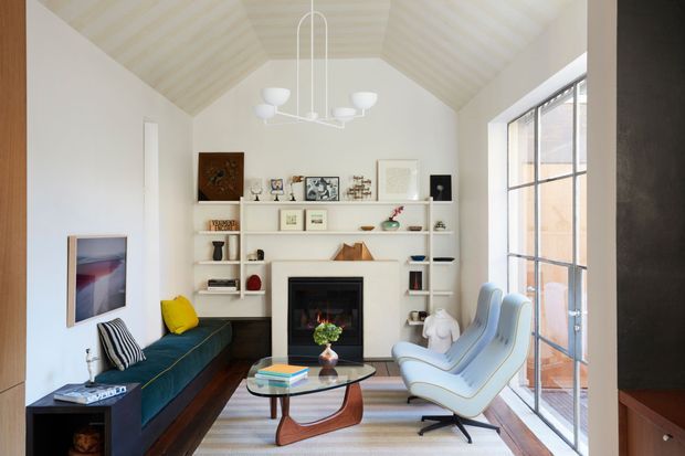 Bright, refined living room with a steep vaulted ceiling, white built-in shelving surrounding a black fireplace, a deep teal sofa, sculptural light-blue lounge chairs, and a glass coffee table. Large black-framed industrial-style glazing opens the room to abundant daylight, while curated artwork and objects add character.