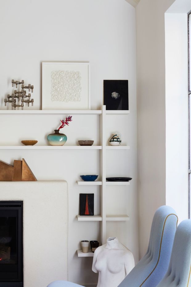 A refined interior vignette showing a white built-in shelving unit surrounding a fireplace, styled with artwork, ceramics, books and sculptural objects. A pale upholstered armchair with contrasting piping sits beside the shelving, while a tall window brings in abundant natural light. The space has a quiet, curated and contemporary character.