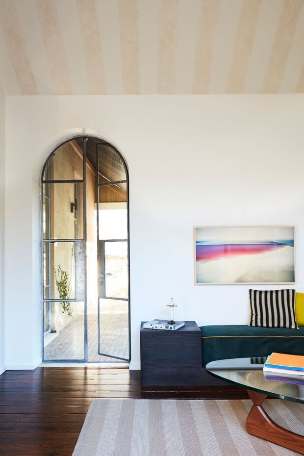 Bright, refined living room with white walls, dark timber flooring, and a high painted ceiling featuring broad exposed-looking timber-toned boards. An arched steel-framed glazed door opens to a timber deck, while a dark teal upholstered sofa, striped cushions, abstract artwork, sculptural coffee table, and pale striped rug furnish the room.