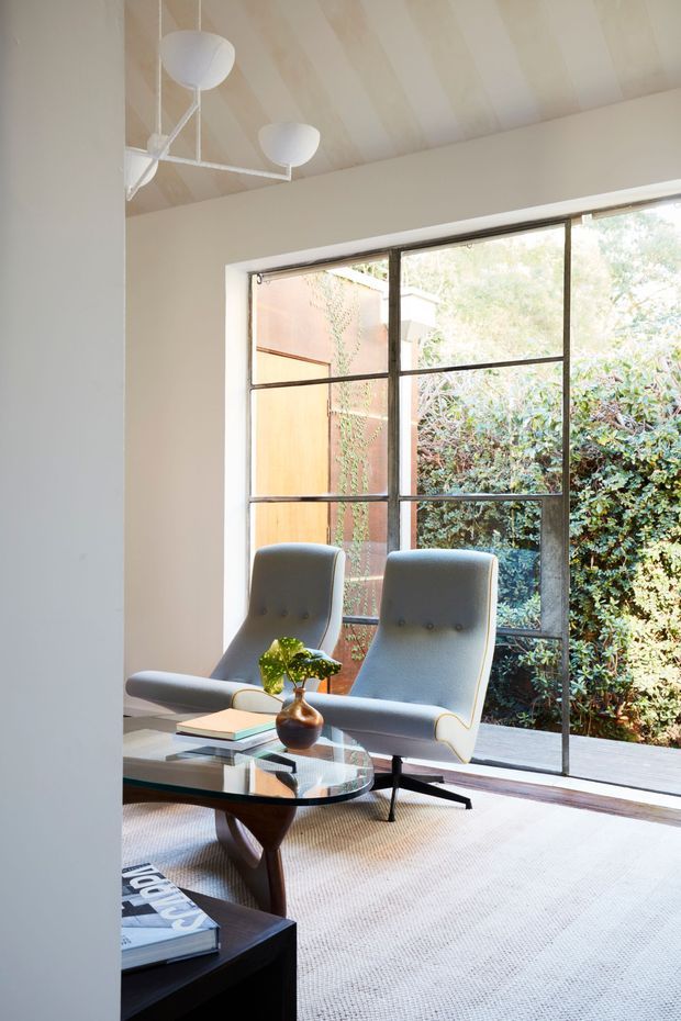 Bright, refined sitting area with two sculptural upholstered lounge chairs arranged around a glass-topped wooden coffee table. A large black-framed grid window overlooks dense greenery and a timber-clad courtyard wall. The room has pale walls, a softly striped vaulted ceiling, a large light rug, and a sculptural white pendant light, creating a calm mid-century-influenced interior.
