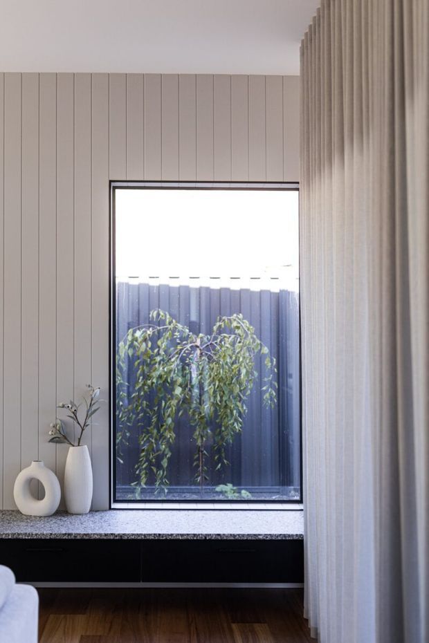 A softly lit contemporary interior featuring a tall black-framed window, vertical light-grey wall panelling, full-height sheer curtains, and a dark built-in cabinet beneath a speckled stone surface. Minimal ceramic vases and branches sit on the ledge, with timber flooring visible below. A small planted courtyard and dark fence are visible through the window.