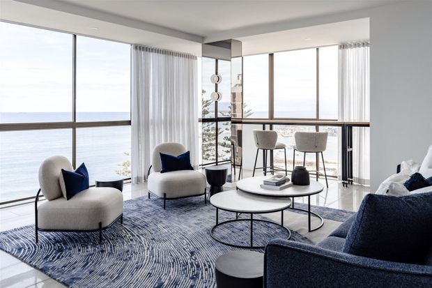 Bright contemporary living room with floor-to-ceiling corner glazing overlooking a calm blue waterfront. The space features pale upholstered lounge chairs, a deep navy sofa, layered round coffee tables, a patterned blue-and-white rug, a slim bar-height counter with stools, sheer curtains, and a tall mirrored or reflective central column. The palette is refined and coastal-leaning, with strong natural light and expansive views.