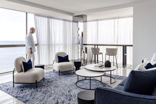 Bright contemporary living room with expansive floor-to-ceiling glazing and sheer curtains overlooking a body of water. The room features sculptural upholstered lounge chairs, a deep blue sofa, layered round coffee tables, a large patterned rug, a mirrored column, and a slim dining or bar area along the window.