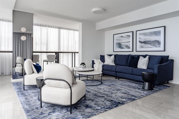 Bright contemporary living room with a deep navy sectional sofa, cream boucle lounge chairs, a round coffee table, and a large abstract blue-and-white rug. Floor-to-ceiling windows with sheer curtains provide abundant natural light, while framed abstract artwork and a sculptural side table add refined accents.