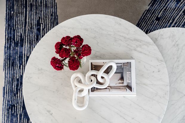 Top-down view of a round white marble coffee table styled with deep red flowers, a white sculptural knot ornament, and a small framed image. The table sits over a neutral grey carpet with bold navy-and-white striped rugs or textile panels visible around it. The composition is decorative and editorial, with no clear room context beyond the styled tabletop.