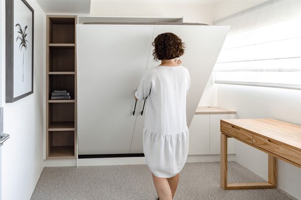 Bright, minimalist home office with a large white concealed storage or wall panel being opened, built-in dark timber shelving, a light timber desk, pale carpet, and a wide horizontal window with translucent blinds. The room has clean contemporary detailing and a neutral palette.