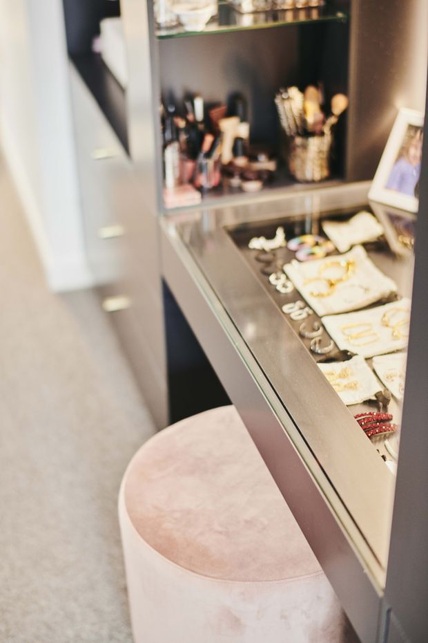 A close-up view of a stylish retail display or dressing area, featuring dark built-in shelving, a glass-front display cabinet filled with jewellery and accessories, makeup brushes and cosmetics, and a round blush-pink upholstered stool. The space has a soft, refined boutique feel with neutral flooring and warm, muted tones.