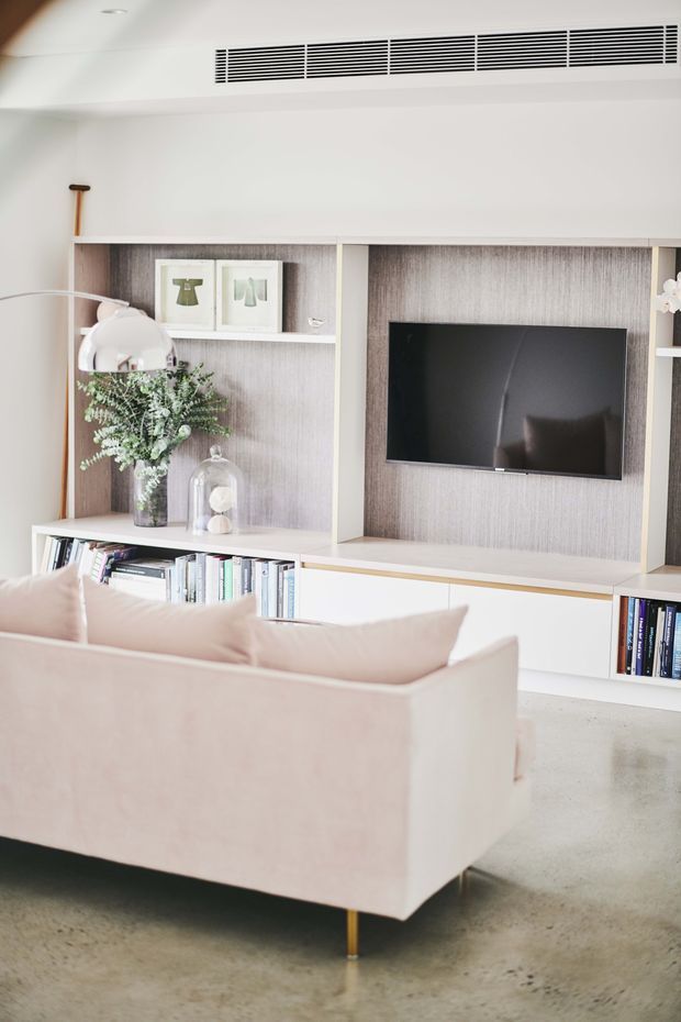 A bright, refined living room featuring a pale blush upholstered sofa in the foreground, a built-in wall unit with pale timber-textured backing, white cabinetry and open shelving, and a wall-mounted television. Decorative framed artwork, books, a glass cloche and greenery sit within the shelving. The room has a polished concrete floor, white walls, a slim floor lamp and linear ceiling air-conditioning vent.