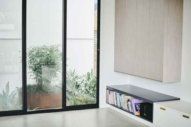 A bright minimalist interior corner featuring full-height black-framed glazing overlooking a planted courtyard, a pale polished concrete floor, and a built-in low storage and bookshelf unit. The joinery combines a dark grey benchtop and open shelving with pale timber wall cabinetry and white drawers with small brass pulls.