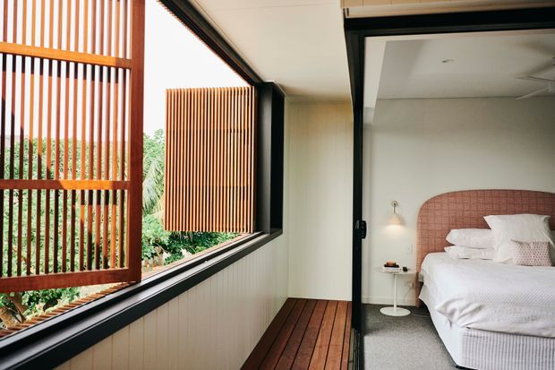 A bright contemporary bedroom viewed through a partially enclosed balcony or sunroom. The room contains a bed with a rounded pink upholstered headboard, white bedding, bedside table and wall light, while the adjacent space features timber decking, white vertical wall cladding, black-framed glazing and prominent vertical timber screening with lush greenery beyond.