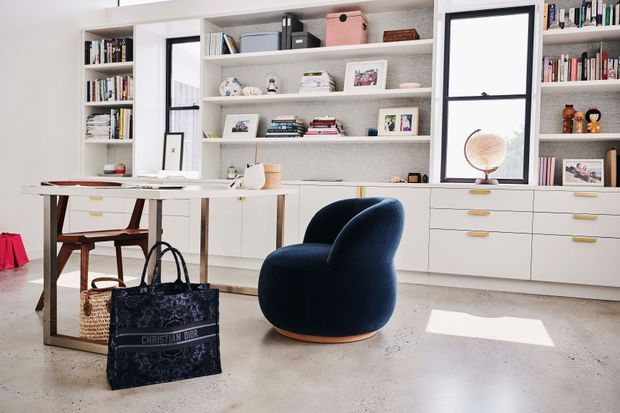 Bright, refined home office with extensive built-in white shelving and cabinetry, a central desk, books and curated display objects, and a sculptural navy lounge chair. Large black-framed windows and a polished light concrete floor add architectural contrast, while natural light creates an airy atmosphere.