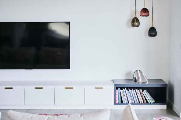 Minimalist contemporary living room media wall with a wall-mounted flat-screen television, long white built-in cabinetry, and a contrasting dark open shelving unit filled with books. Three sculptural pendant lights hang at different heights, and a metallic decorative object sits on the shelving. The space has a clean, restrained palette and low-profile built-in joinery.