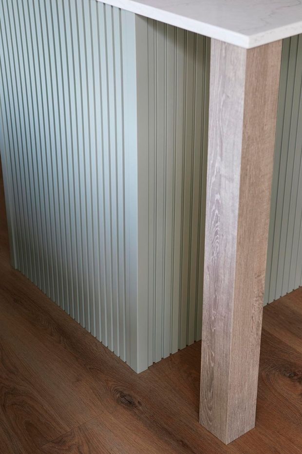 Close-up interior detail of a fluted, vertically ribbed muted sage-green island or built-in joinery, paired with a pale stone-look benchtop and a light timber support panel. The space has warm brown timber flooring and a restrained contemporary palette.