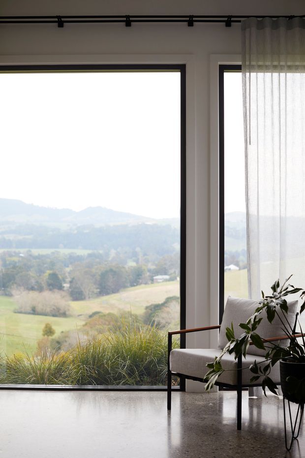 A calm, light-filled interior with expansive black-framed floor-to-ceiling glazing overlooking rolling rural hills. A minimalist upholstered armchair, sheer curtains, potted plant, and indoor grasses sit against a polished concrete floor, creating a strong connection to the landscape.