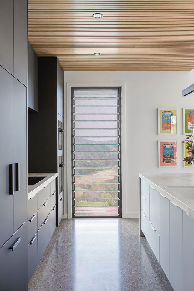 A bright, contemporary galley kitchen with full-height dark grey cabinetry on the left and a white island with a pale stone benchtop on the right. A full-height louvred window or glazed door provides filtered natural light at the end of the room. The ceiling is lined with narrow light timber battens and recessed downlights, while the polished terrazzo-style floor adds a speckled finish. Framed artwork is displayed on the right wall.