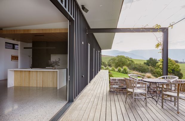 A contemporary indoor-outdoor living area opening onto a broad timber deck. The interior includes a compact kitchen with a white island, vertical timber detailing, dark cabinetry or wall panelling, and a polished speckled floor. Large dark-framed glazed openings connect it to the deck, which has an outdoor dining setting, brick-edged planter, pergola structure, and views toward rolling green hills. The design has a clean, integrated indoor-outdoor character with strong linear detailing.