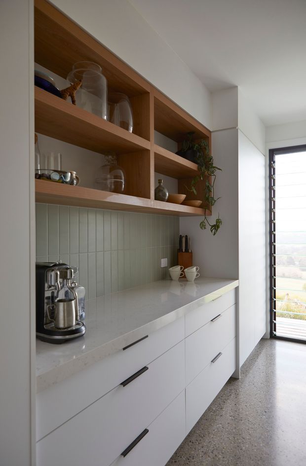 A refined built-in kitchen or beverage station with white base cabinetry, a pale stone benchtop, muted green vertical tile splashback, and open timber display shelving. The shelves hold glassware, ceramics and plants, while a coffee machine and cups sit on the counter. A tall louvred window at the end brings in natural light, and the floor is finished in terrazzo or polished aggregate.