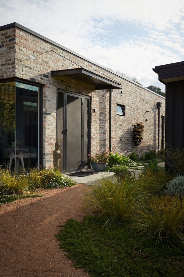 Exterior view of a contemporary single-storey building with a long pale exposed-brick facade, dark metal roof edging, black-framed glazing and a sheltered entry door. A gravel garden path leads through low ornamental grasses and planted beds toward the entrance. A dark-clad adjoining volume is visible at right.