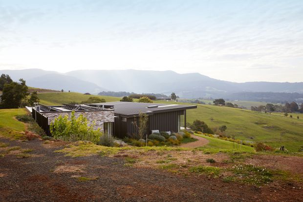 A low-profile contemporary rural house set into a gently sloping green landscape. The building has dark vertical cladding, a broad flat roof, extensive glazed openings, and a partially enclosed outdoor area with a lightweight slatted roof structure. Rolling farmland and layered mountain ranges form the background.
