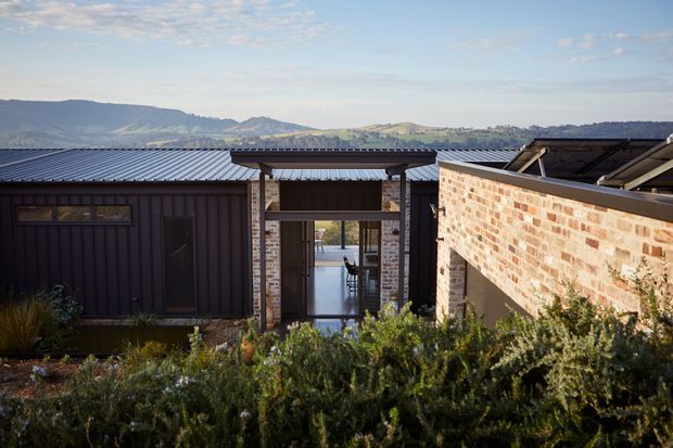Exterior view of a low-rise contemporary building set against rolling hills, with dark vertically ribbed metal cladding, corrugated metal roofs, exposed brick walls, and a covered glazed entry or courtyard connection. Native-looking landscaping fills the foreground, while a glimpse of an interior dining or living space is visible through the central opening.