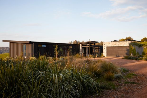 Single-storey contemporary residence viewed from the landscaped approach, with a long low dark-clad building, broad shallow rooflines, a partially sheltered entry or outdoor living zone, and a separate light masonry garage. Native grasses and drought-tolerant planting surround the gravel driveway and house. The setting appears semi-rural or bush-adjacent, with distant vegetation visible beyond the property.
