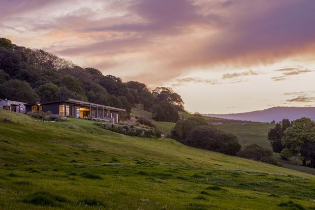 A low, elongated contemporary house set into a broad, gently sloping rural landscape. The building features dark horizontal cladding, extensive floor-to-ceiling glazing, and a projecting roofline, with landscaped planting around the partially embedded structure. Rolling pasture, wooded hills, and a dramatic warm purple-pink sunset dominate the setting.
