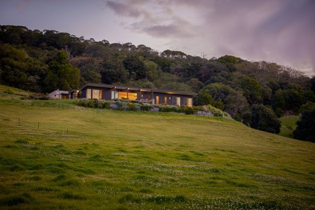 A low, modern single-storey house sits within a broad grassy rural clearing at dusk, backed by dense native-looking woodland. The dwelling has dark horizontal cladding, extensive warm-lit glazing, and a simple low-pitched roofline, with a small detached or adjoining outbuilding to the left. The scene has a secluded, semi-rural character and subdued evening lighting.