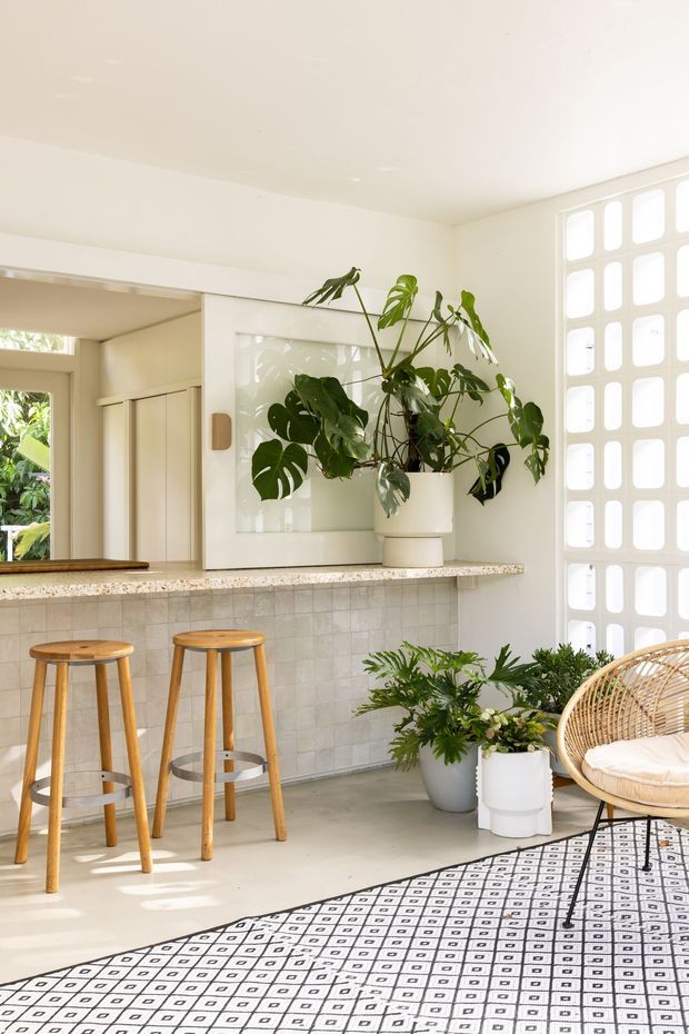 Bright, plant-filled open-plan kitchen and living area with a tiled kitchen island, terrazzo benchtop, timber bar stools, patterned rug, rattan lounge chair, and a white breeze-block feature wall. The space has abundant natural light and a relaxed indoor-outdoor feel.