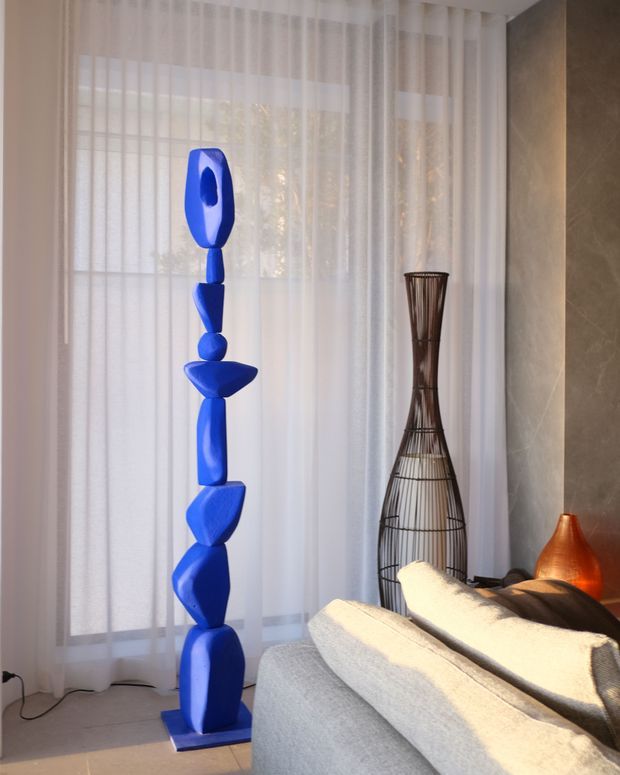A refined contemporary living room corner featuring a tall cobalt-blue sculptural artwork beside sheer floor-to-ceiling curtains. A pale upholstered sofa occupies the foreground, with a tall dark woven sculptural vase and a warm copper-orange lamp against a textured stone or plaster feature wall. Soft daylight creates a calm, gallery-like atmosphere.